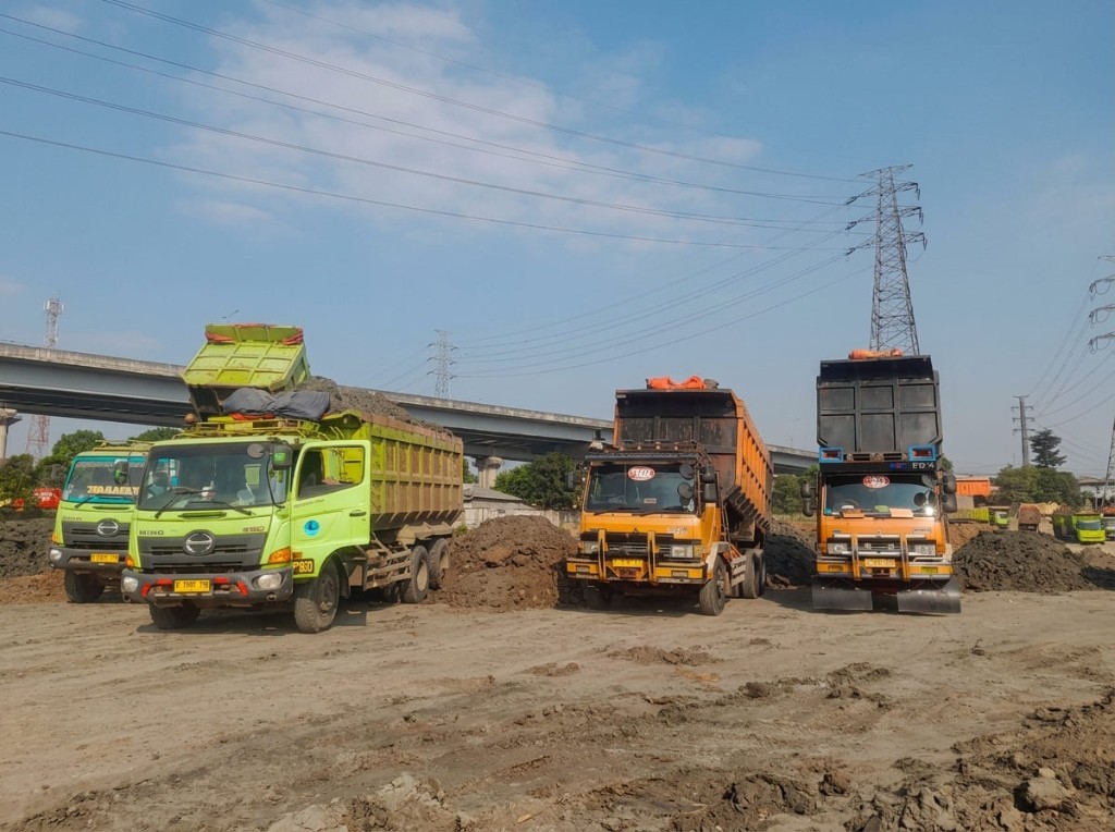 Dump truck antre di area timbunan, dengan jalan layang di belakang.