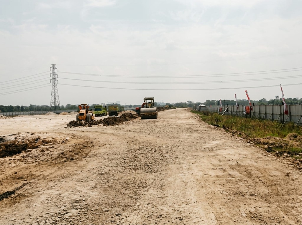 Pemadatan jalan tanah di kawasan proyek, dengan roller dan armada di kejauhan.